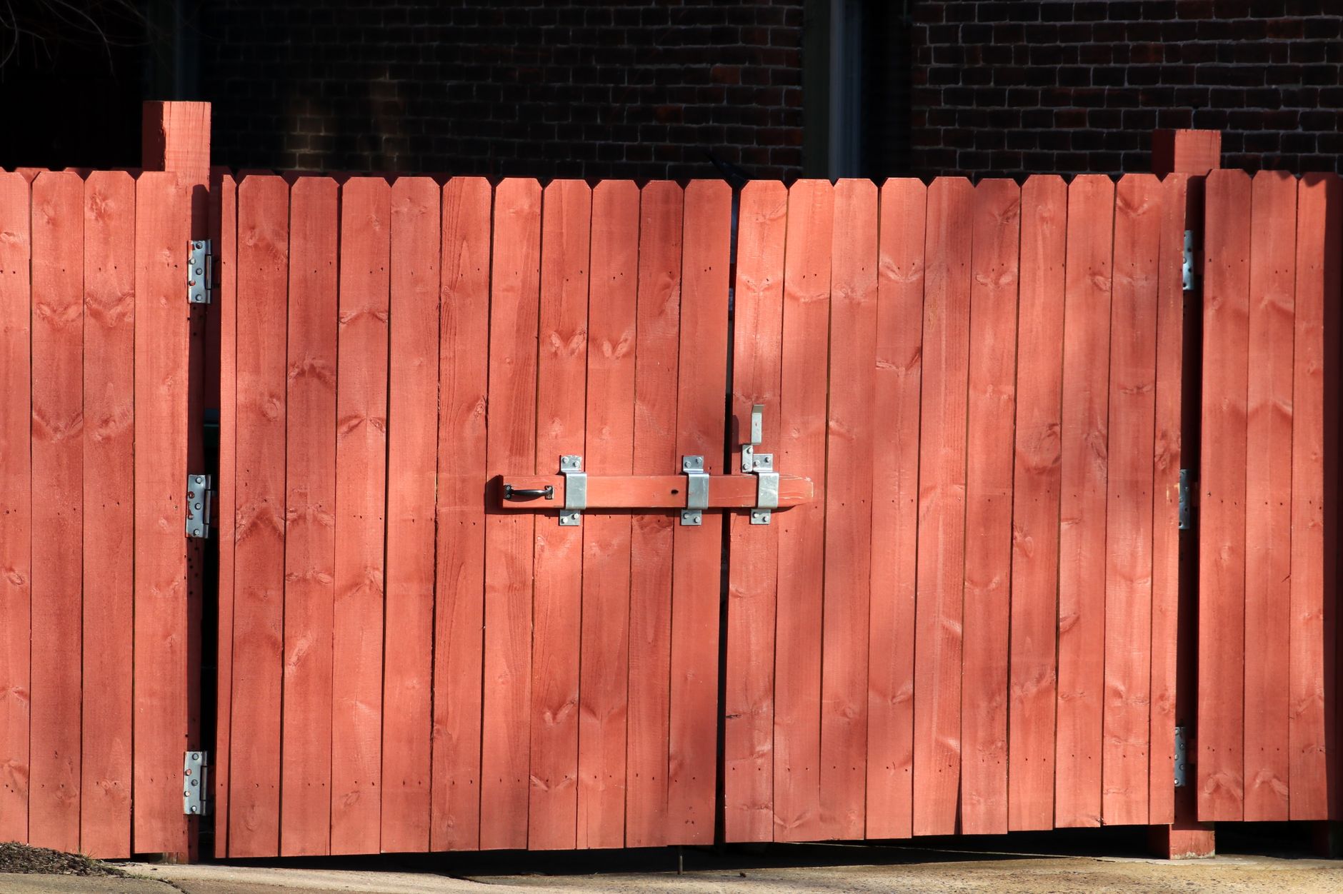 A closed wooden fence gate with a latch and hinges, set against a brick wall background.